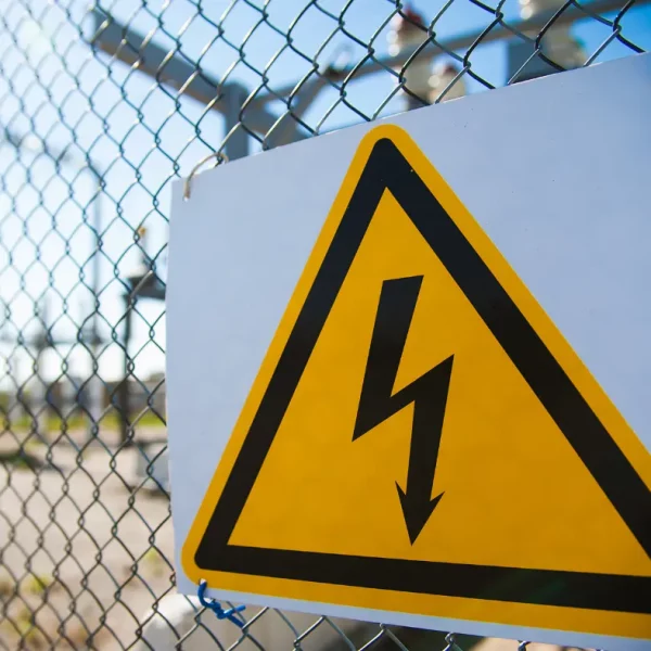 Panneau d'avertissement électrique fixé à une clôture métallique dans une zone industrielle, représentant un danger de haute tension. Control Safe Energy.